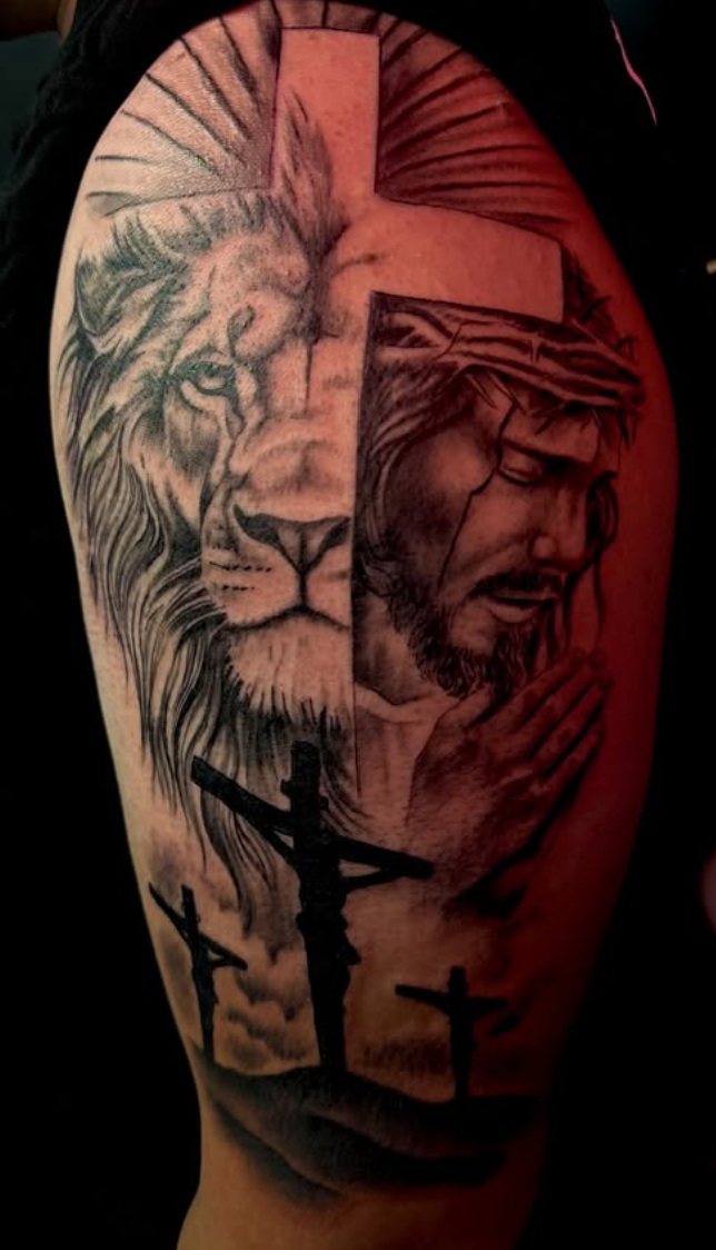 Black-and-grey realism tattoo of a lion split with a praying figure and crosses, on the upper arm.