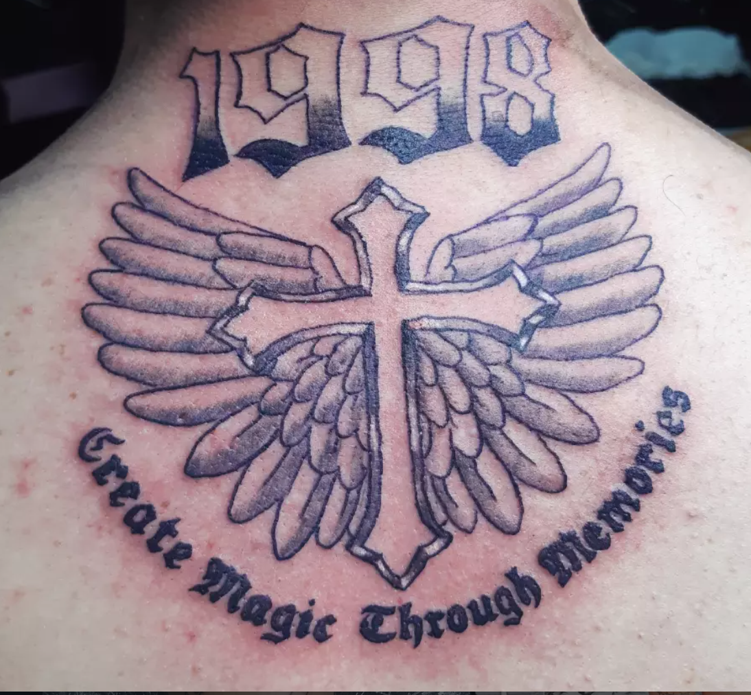 Bold traditional wings and cross memorial tattoo with gothic lettering across the upper back.