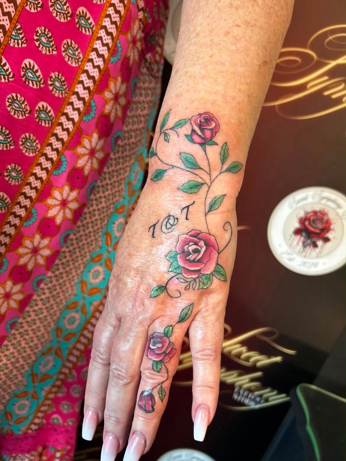 Colour rose vine tattoo winding across the hand and finger with initials.