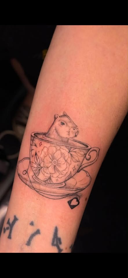Fine-line illustrative capybara sitting in a floral teacup tattoo on the forearm.