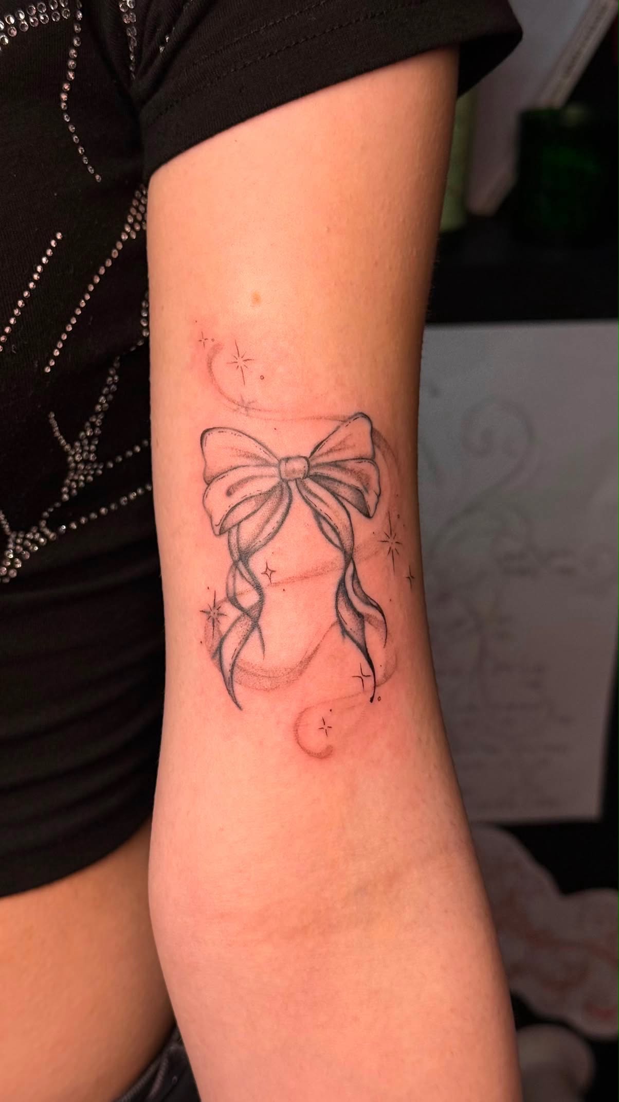 Fine-line bow tattoo with scattered stars on the upper arm.