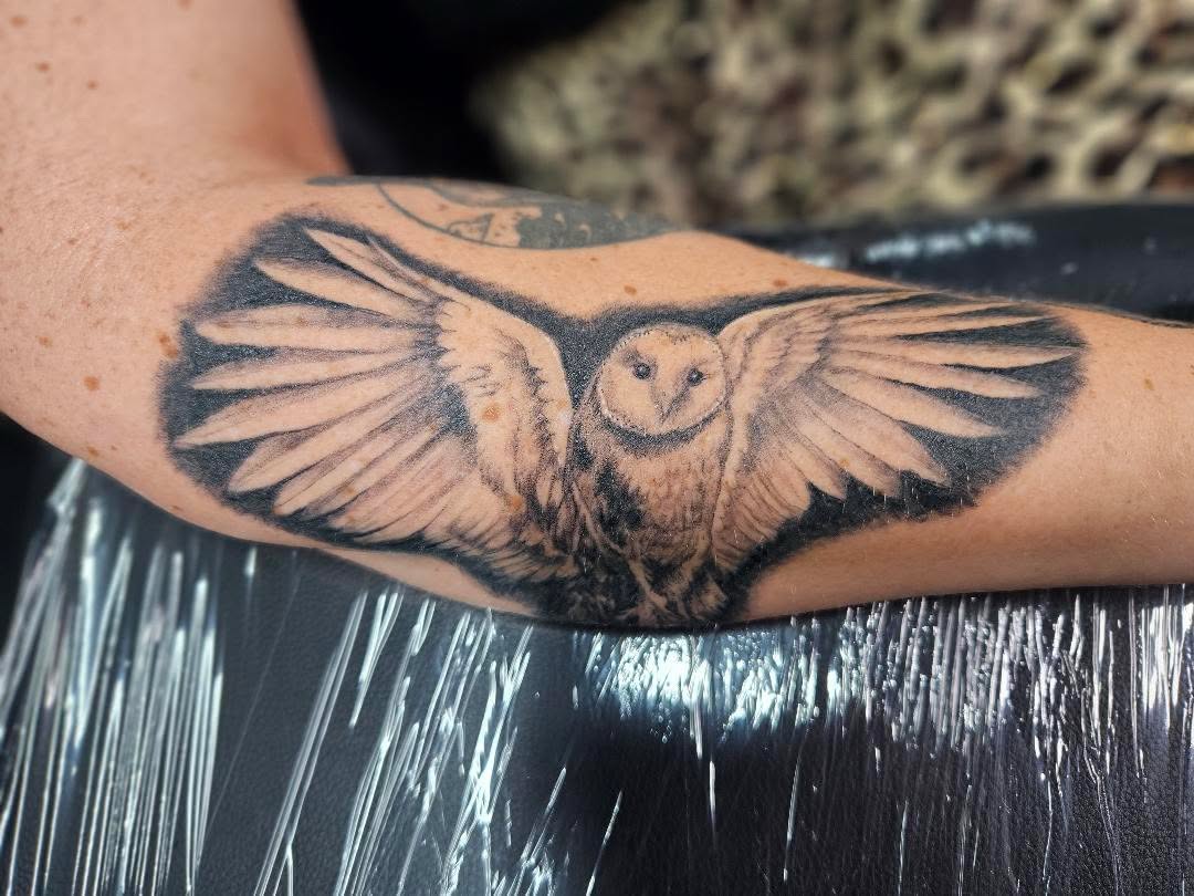 Black-and-grey realistic owl tattoo with its wings spread across the forearm.